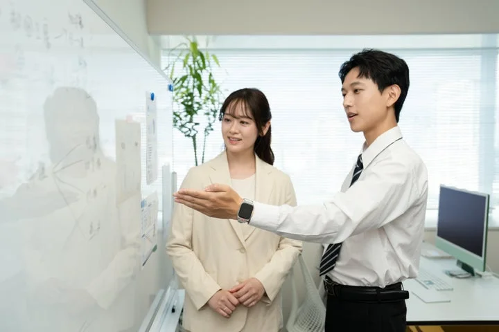 ホワイトボードの前で説明する男性と、それを見守る女性のオフィスシーン