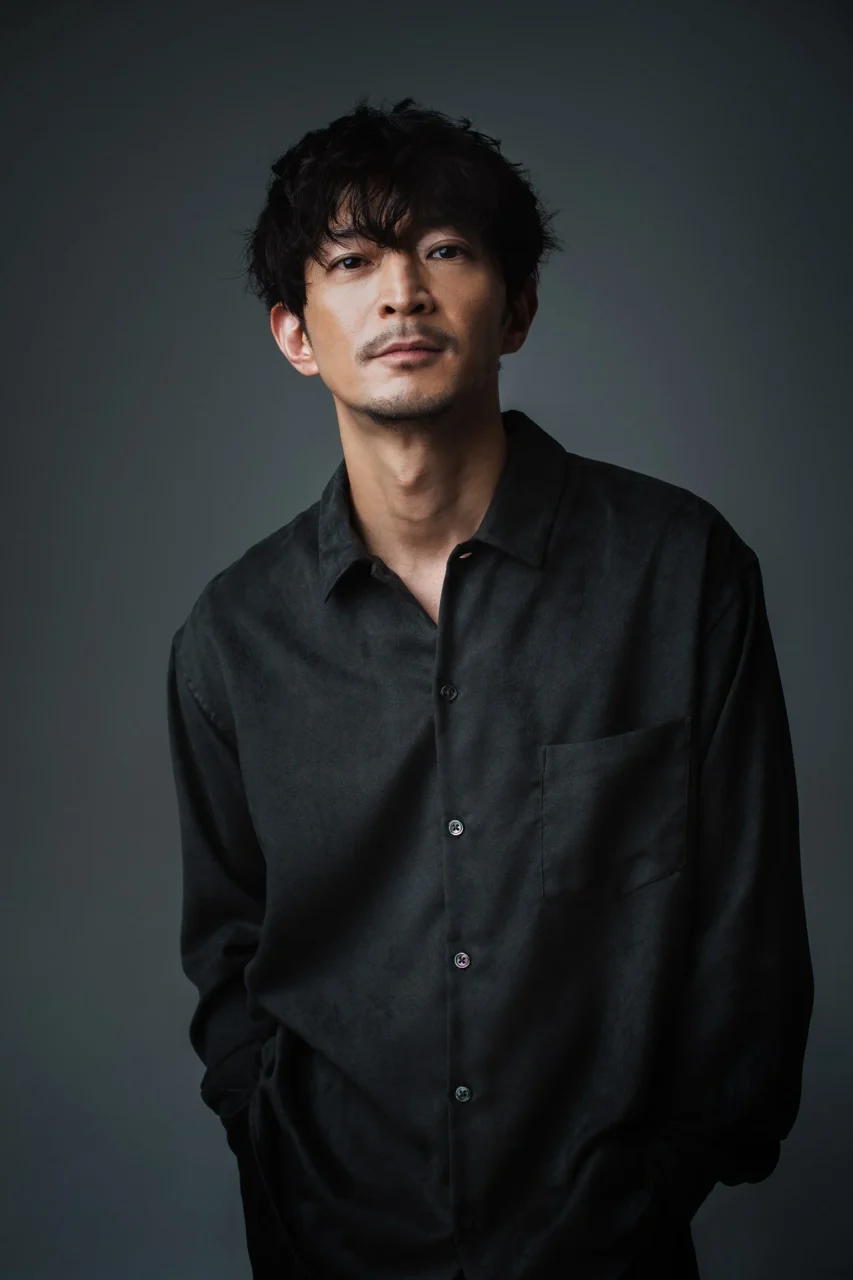 津田健次郎さんのポートレート。黒いシャツを着用し、暗い背景で正面を向いている。