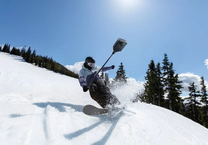 雪山でスノーボードを滑る人物が、セルフィースティックに装着したカメラで撮影している様子