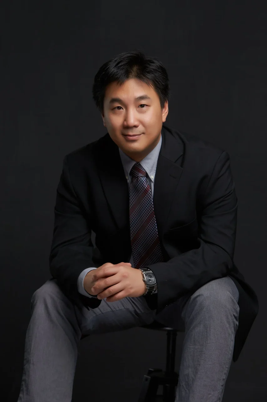 色彩専門家Chris Bai氏のポートレート写真。黒いスーツにネクタイを着用し、椅子に座っている。