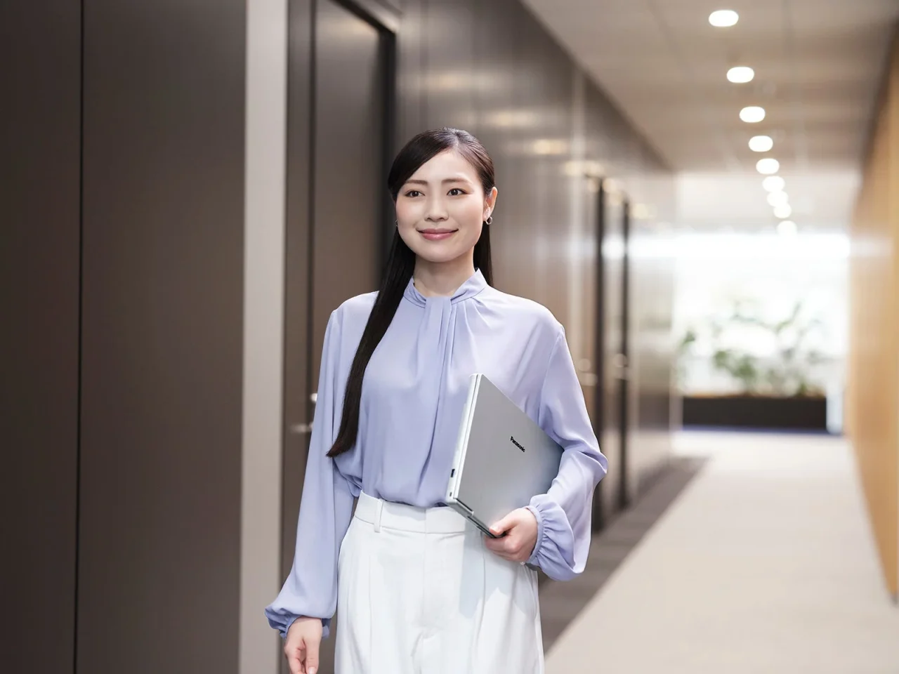 銀色のノートPCを抱えて廊下に立つ、薄紫のブラウスの女性