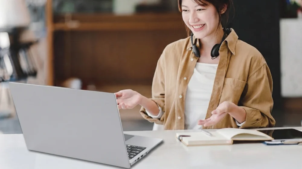 ヘッドセットを首にかけた女性がノートPCに向かい笑顔で話す様子