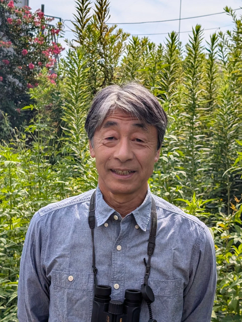 双眼鏡を首にかけ緑の植物の前で微笑む野鳥写真家の中野泰敬氏