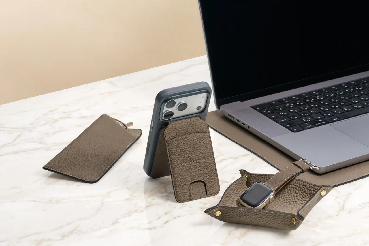 大理石の机に置かれたレザーのカードケース付きiPhone、ノートPC、小物