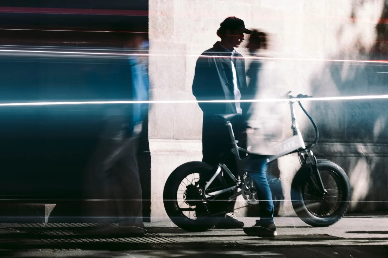 街角で帽子の人物が小型のMATE.BIKEのe-BIKEの横に立つ、光の筋が走る写真