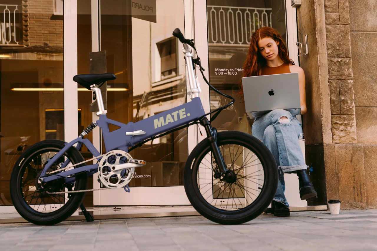 MATE Fusion 2.0。店先に停めた青紫のMATE.の電動自転車と、ノートPCを使う女性