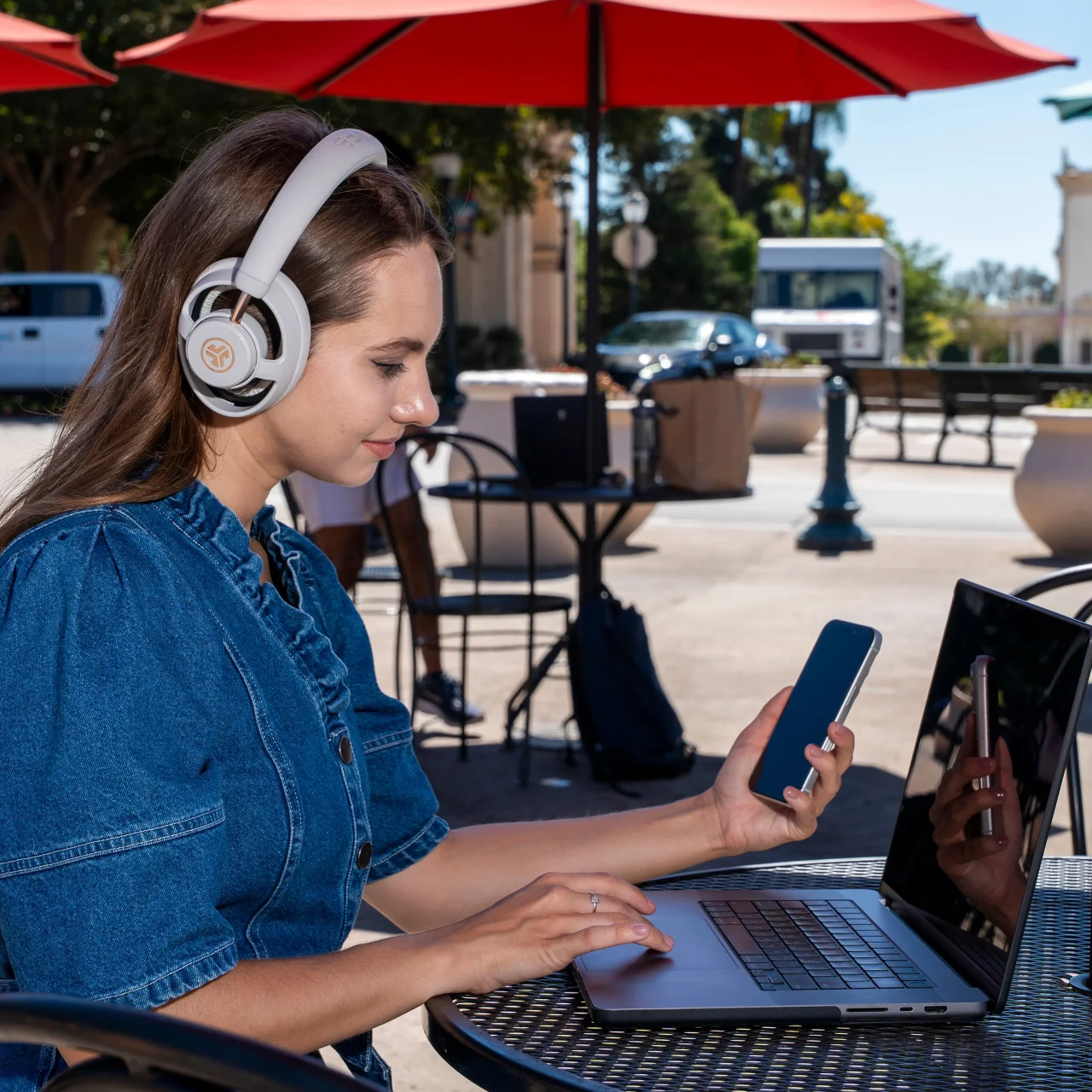 JBuds Open Wireless を着けた女性が屋外でスマホとノートPCを使う様子