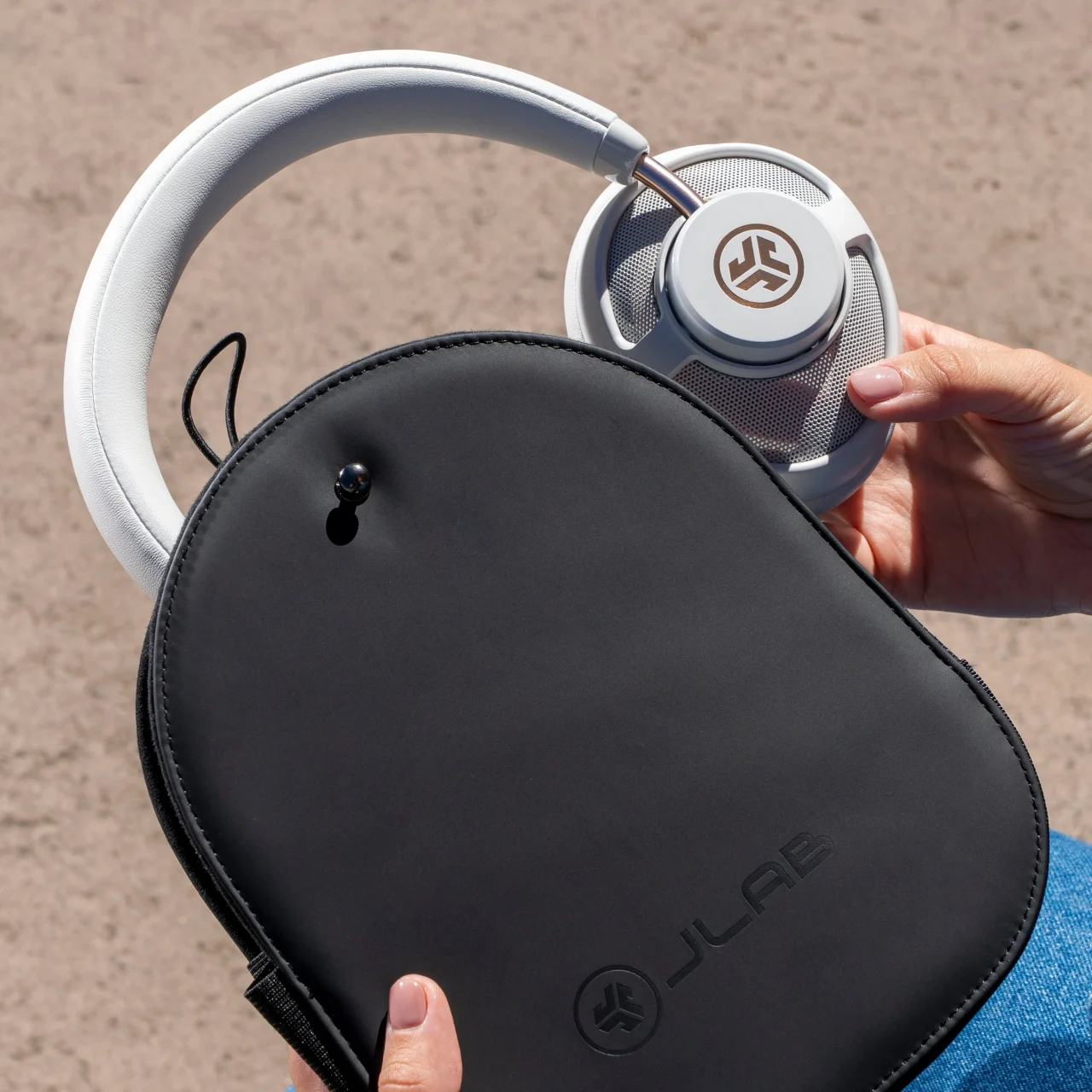 JBuds Open Wireless。白いヘッドホンと黒いJLabロゴ入りケースを持つ手