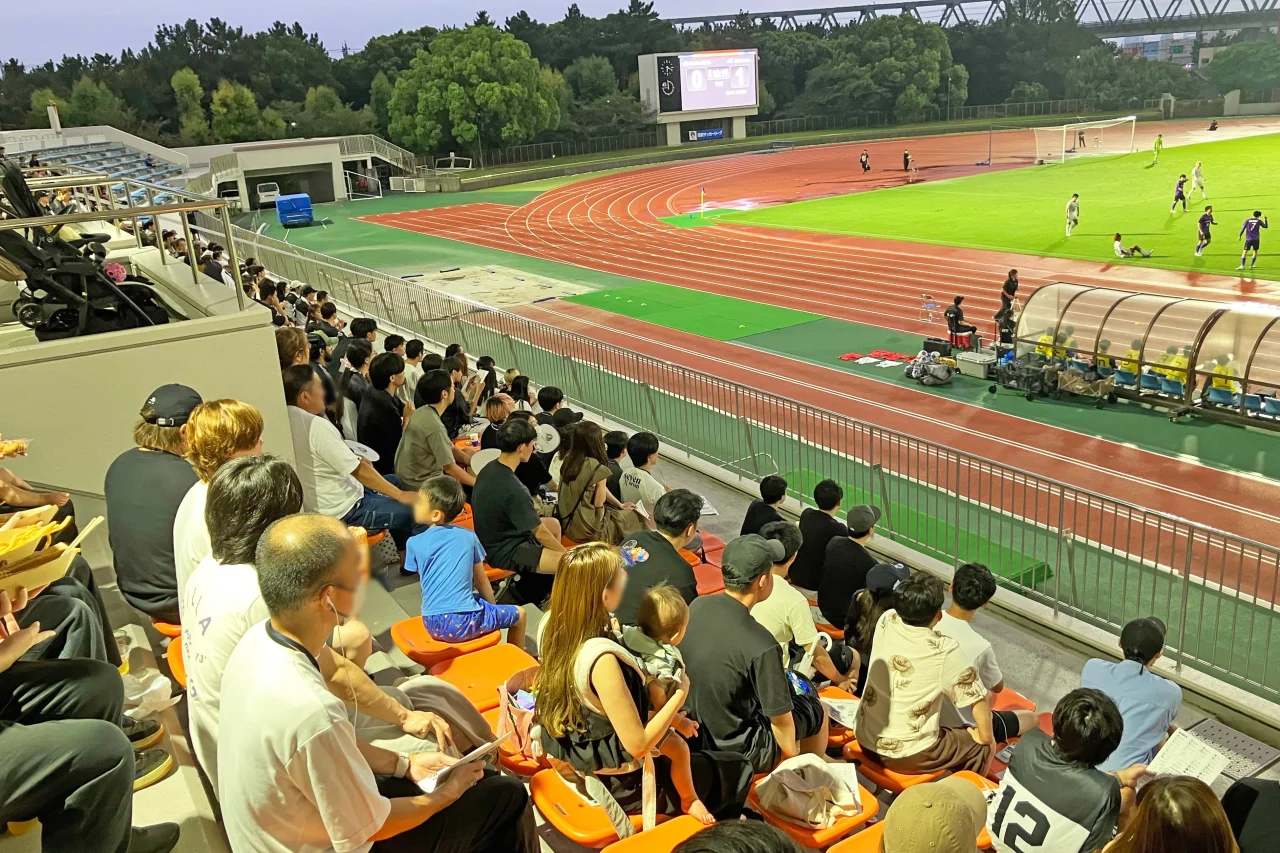 スタジアムの観客席からピッチの試合を観戦する観客たち