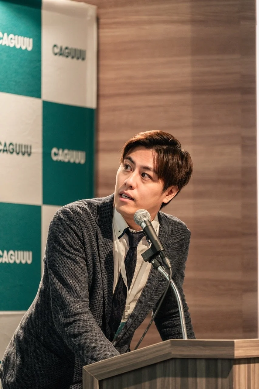 CAGUUU商品開発責任者・速水康博が演台のマイクで登壇する様子