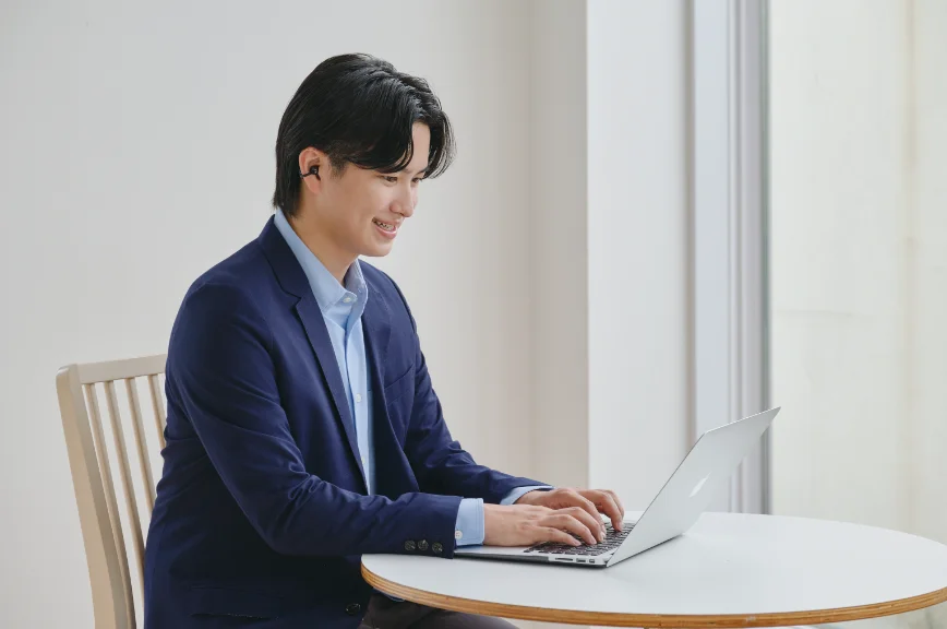 イヤホンを片耳に着けノートPCを操作する紺ジャケットの男性