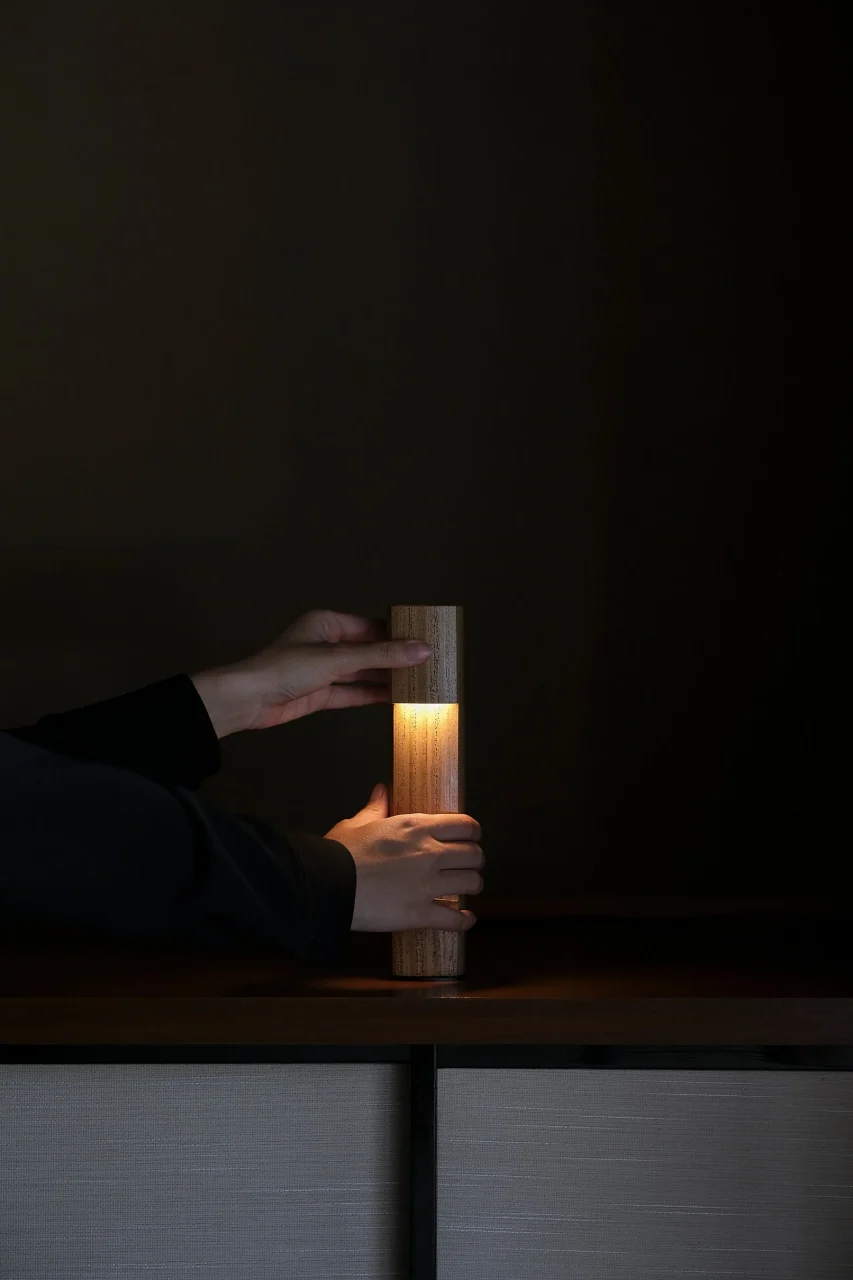 暗い部屋で両手に挟まれ光る、竹製の円柱形照明Nosou 0°
