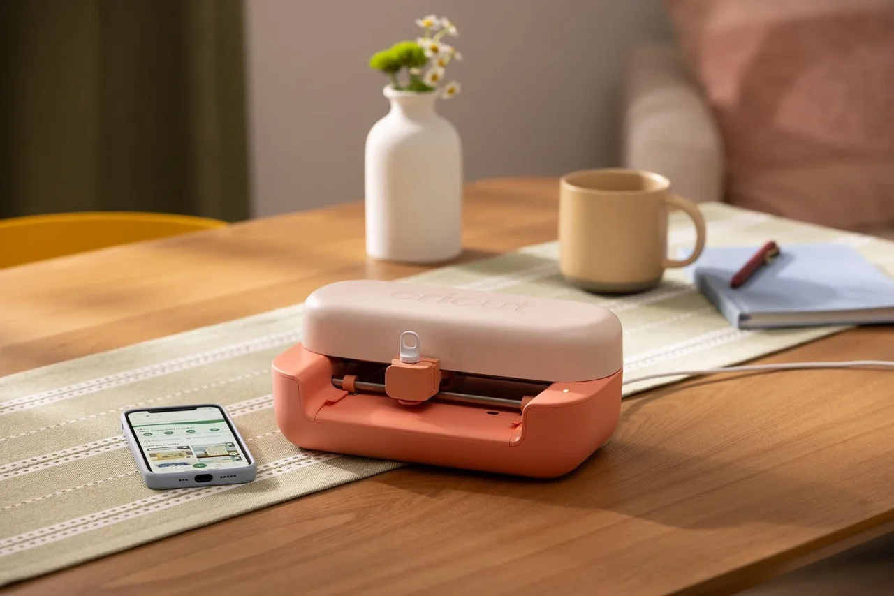 木製テーブルに置かれたコーラル色のCricut Joy 2本体とスマートフォン