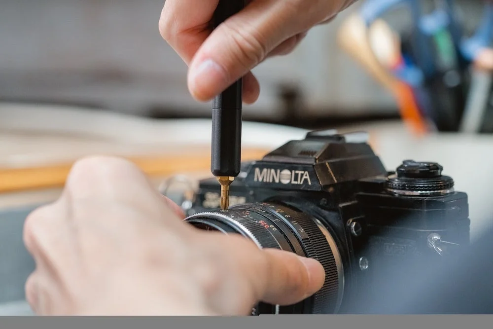 MINOLTAのカメラのレンズ部分を黒いドライバーで作業する手元