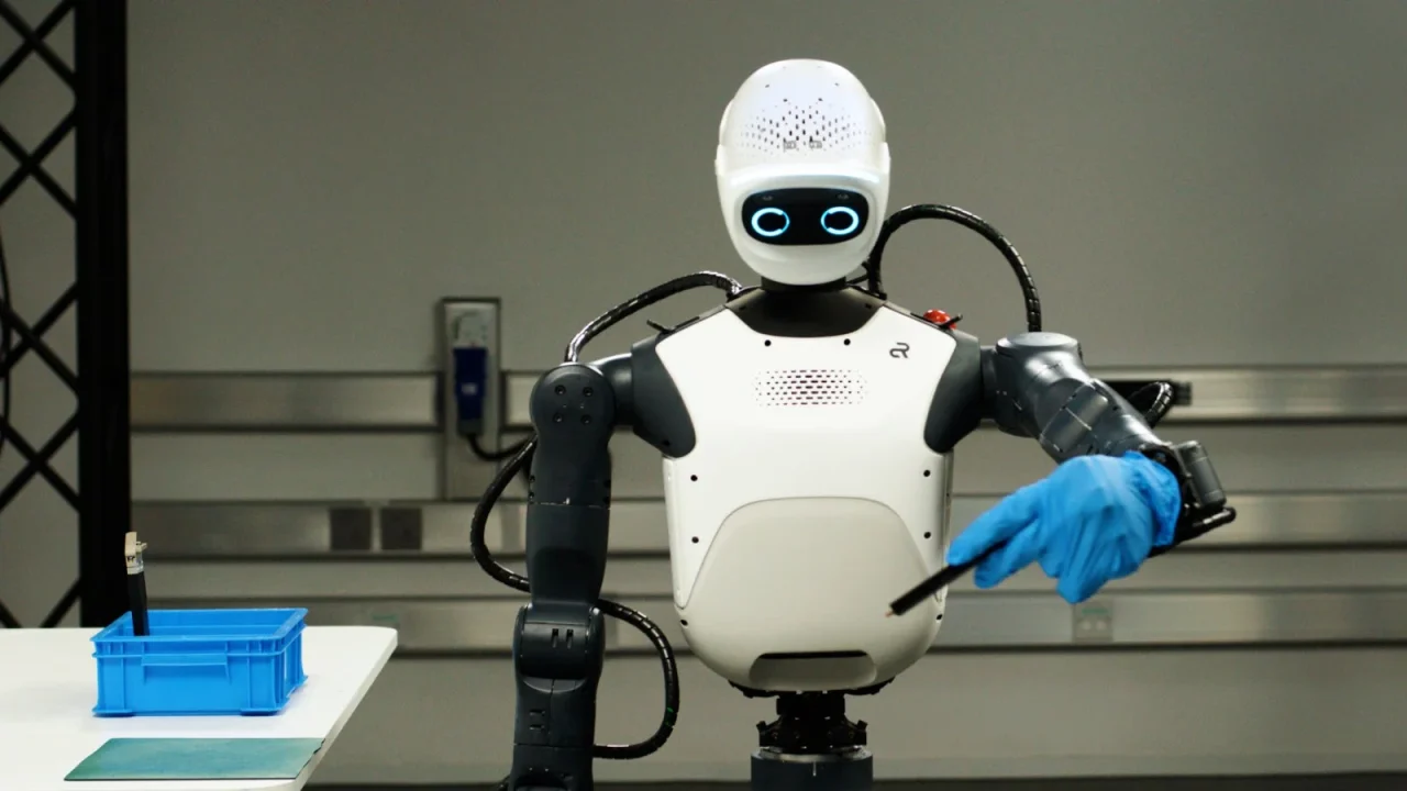 白い頭部と胴体に青い手袋をつけたヒューマノイドロボットが立つ様子