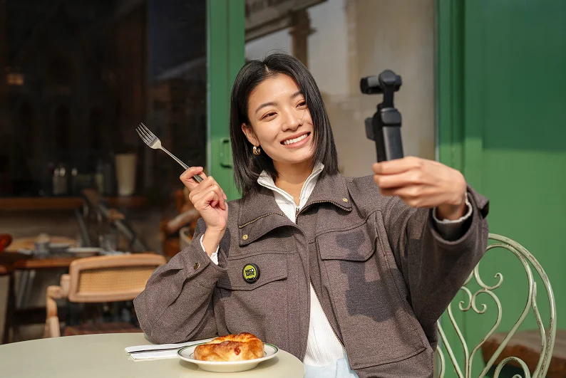 カフェでフォークを持ち、小型カメラを自撮りの向きに構える女性