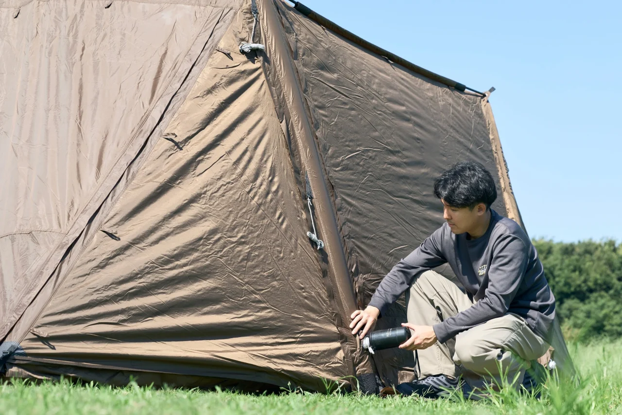 茶色のテントのそばで黒い筒状のAIR TENT PUMPを手に屈む男性