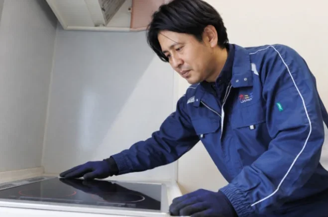 青い作業服と手袋の男性がIHクッキングヒーターの天板に手を添え確認している