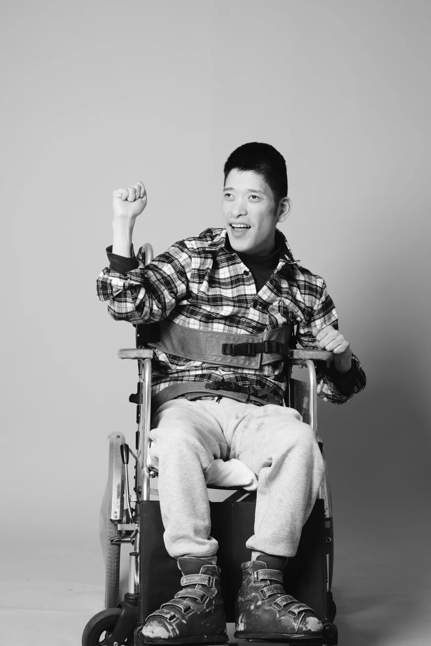 作家 鈴木広大さんの白黒ポートレート。車いすで拳を上げ笑顔