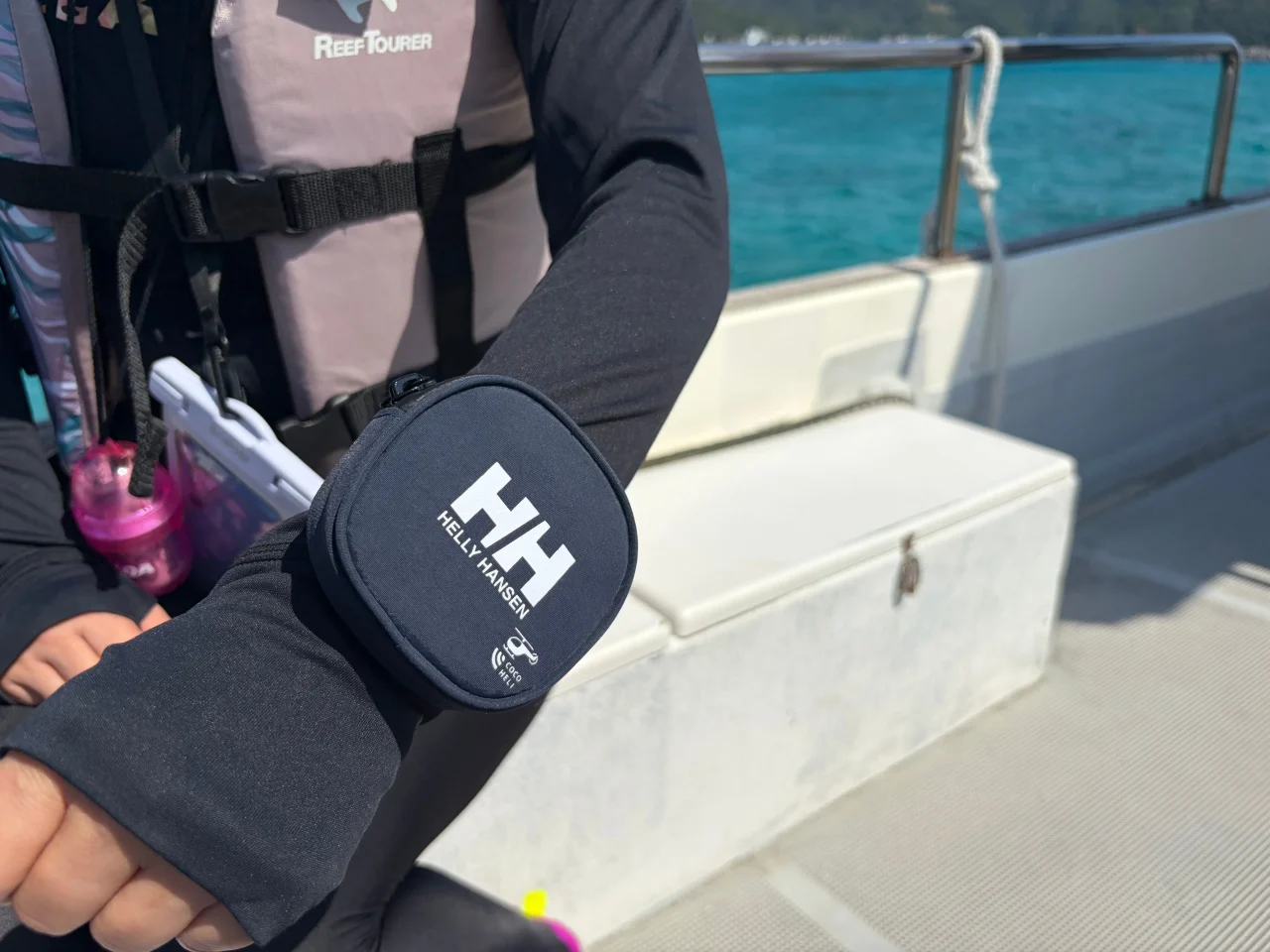 腕に着けた、HELLY HANSENロゴ入り紺色の携行ポーチ型ココヘリマリン発信機
