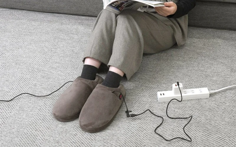 床に座り、黒いケーブル付きのどこでも床暖スリッパを履いて電源タップに接続している様子