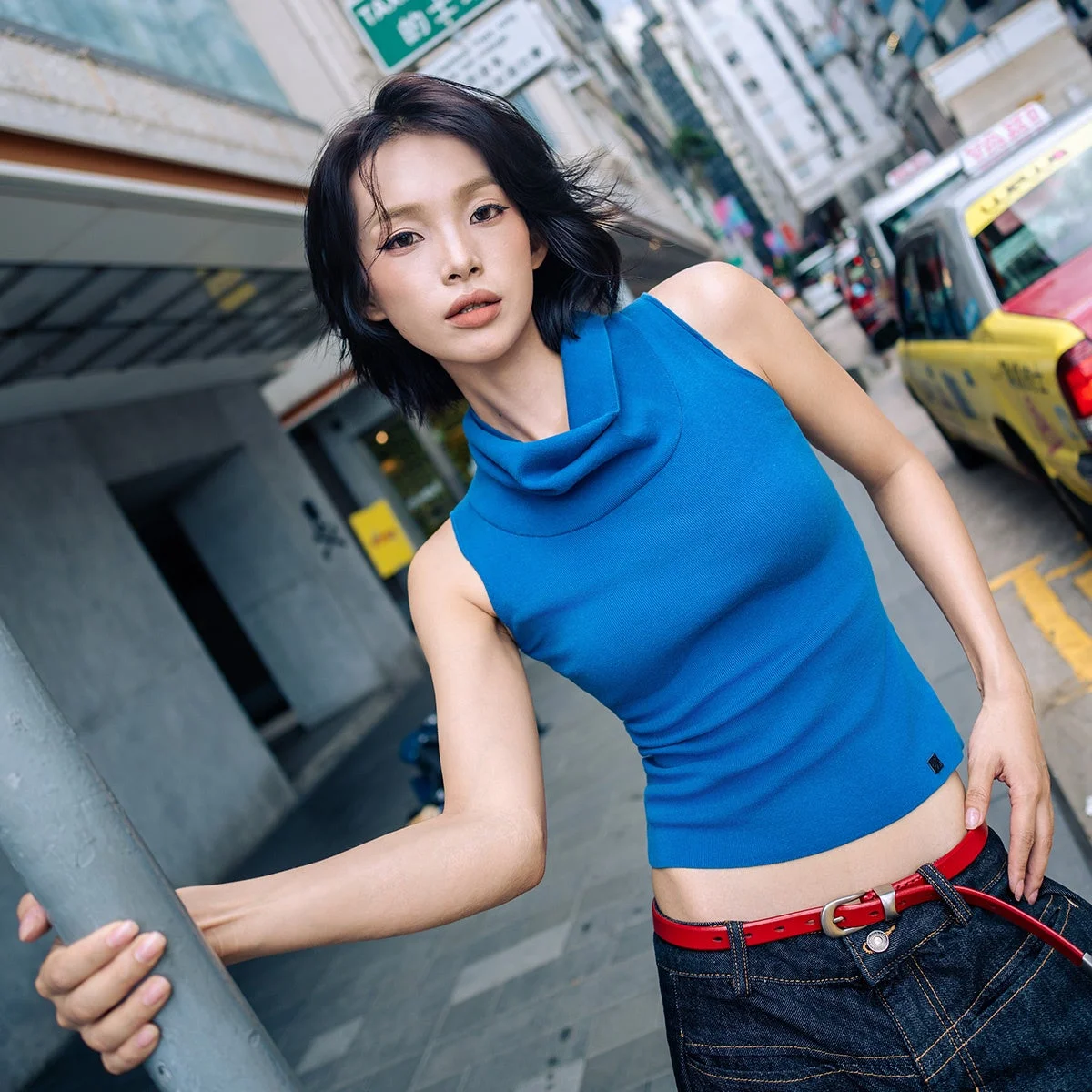 青いハイネックのノースリーブを着て街中に立つ短髪の女性