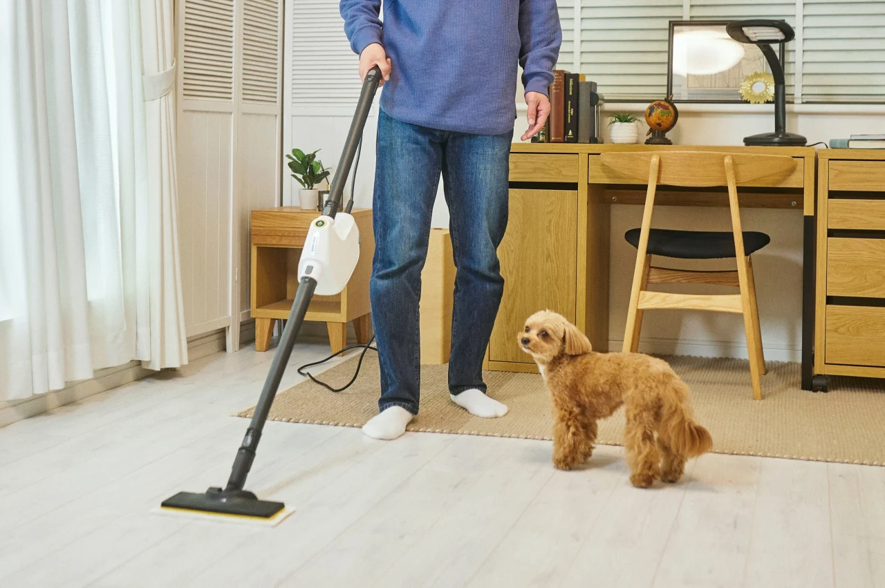 リビングでフローリングを床用ノズルのスチームクリーナーで掃除する人と犬